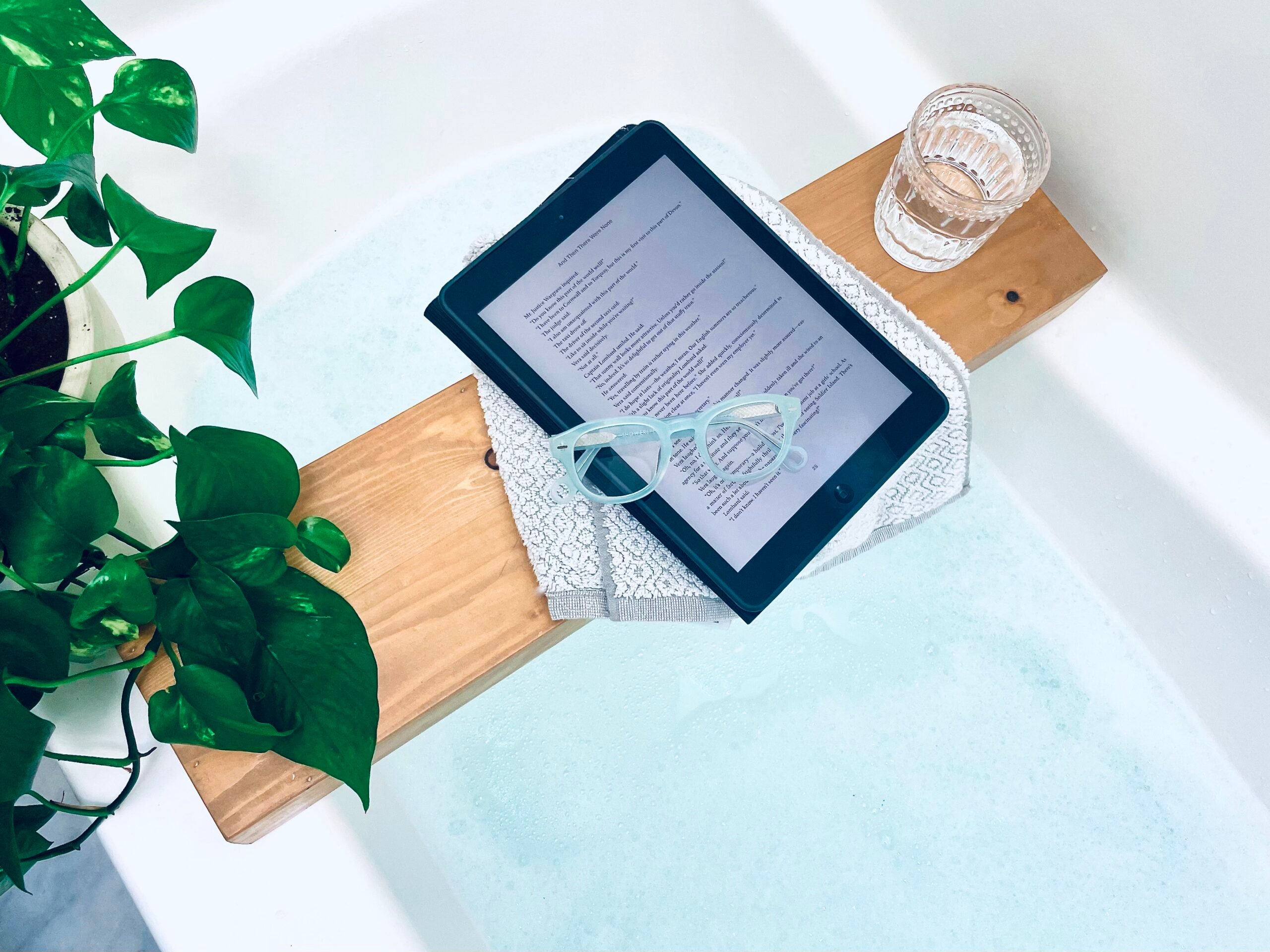 Misty Green Showerspecs reading glasses on a bathtub tray beside a tablet.